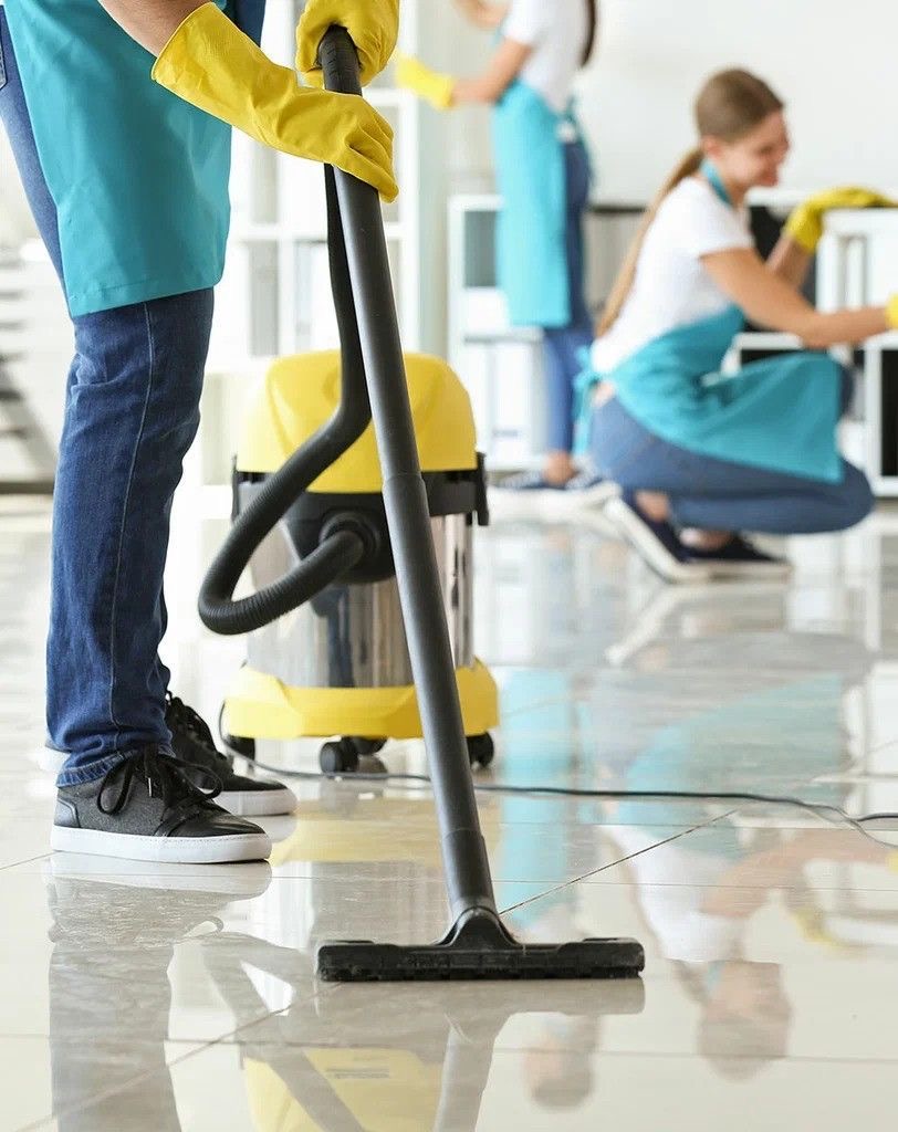 A man and a woman are cleaning a bright, modern room. The woman, wearing pink gloves, is mopping the floor, while the man, wearing yellow gloves, is arranging items on a shelf. They are both dressed in blue overalls and appear focused on their tasks. The room has plants and a colorful abstract painting on the wall.