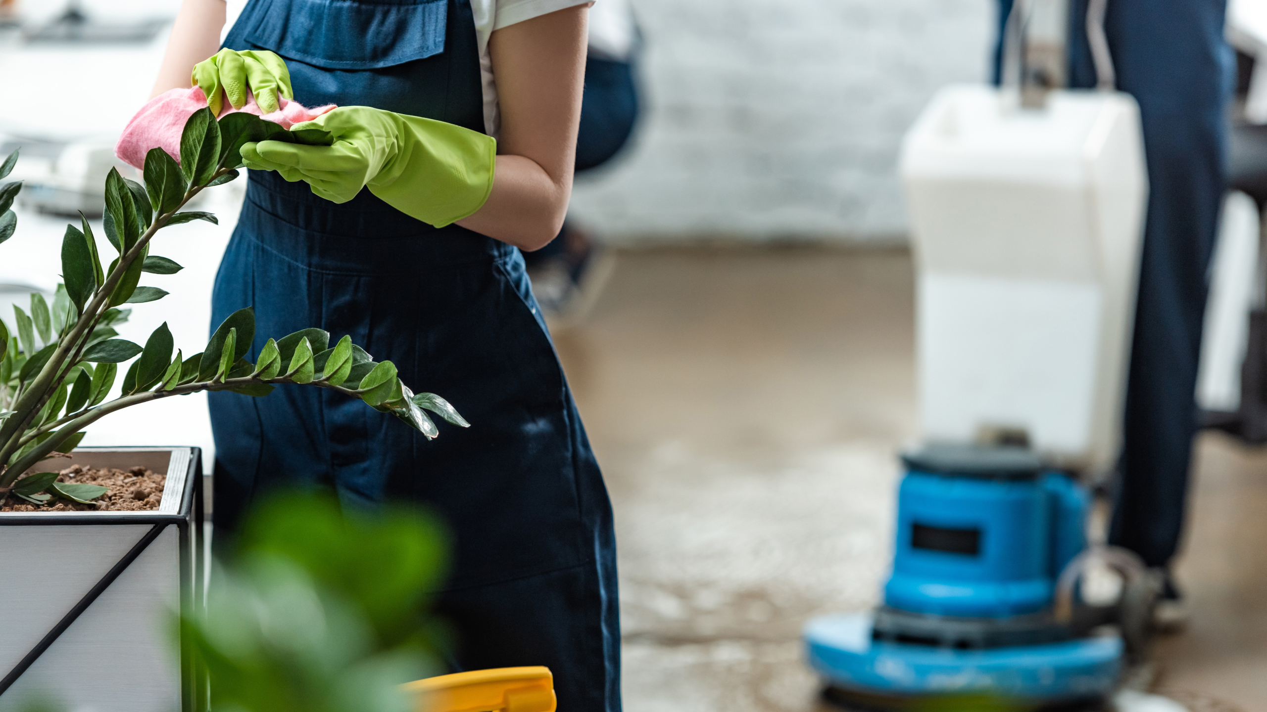 A person wearing green gloves is tending to a plant in a pot, while a blue cleaning machine is visible in the background on a floor.